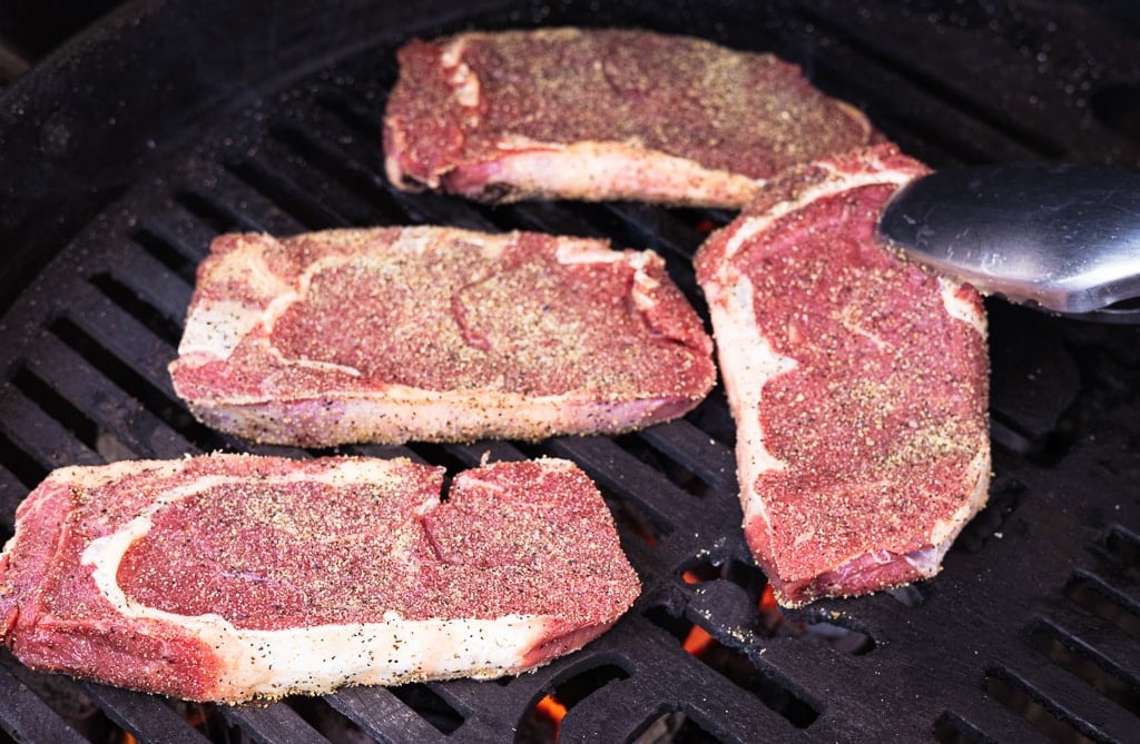 Four seasoned bison ribeye steaks on the grill grates of a charcoal grill.