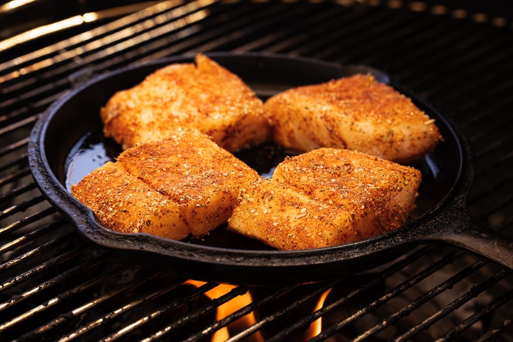 Four seasoned cod fillets in a cast iron skillet on the grill.