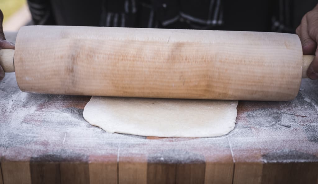 Flour tortilla dough being flattened by a wooden rolling pin.
