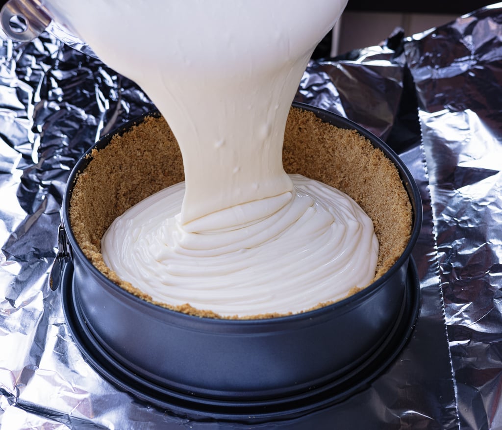 Cheesecake batter pouring into a springform pan lined with graham cracker crust.