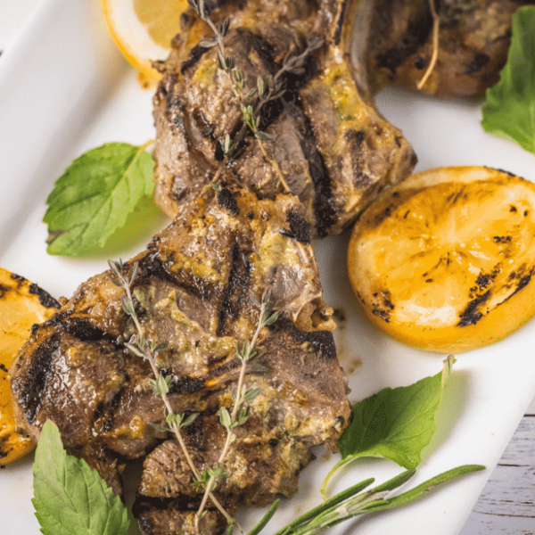 Three grilled lamb chops surrounded by fresh herbs and sliced lemons.