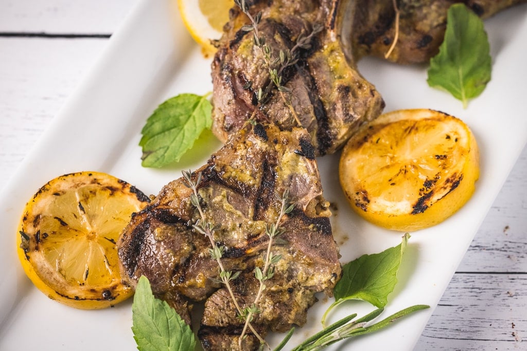 Three grilled lamb chops surrounded by fresh herbs and sliced lemons.