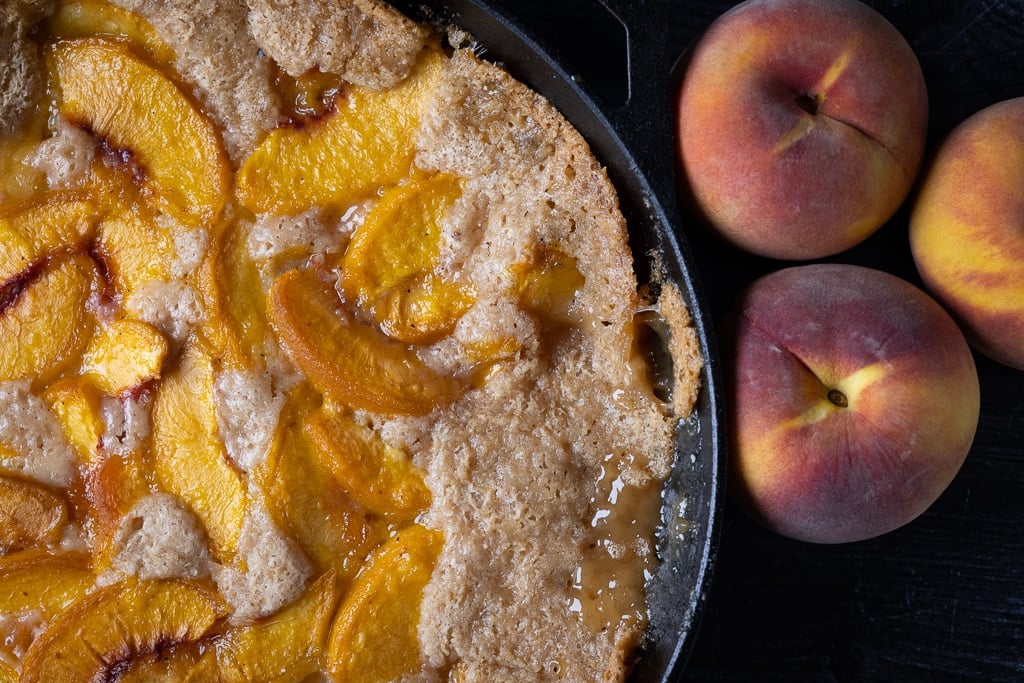 Smoked peach cobbler in a cast iron skillet next to whole fresh peaches.