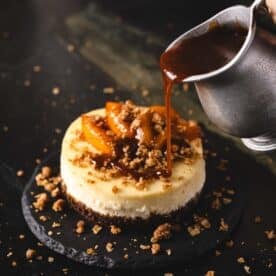 Caramel sauce pouring from a metal pitcher onto a cheesecake topped with peaches.
