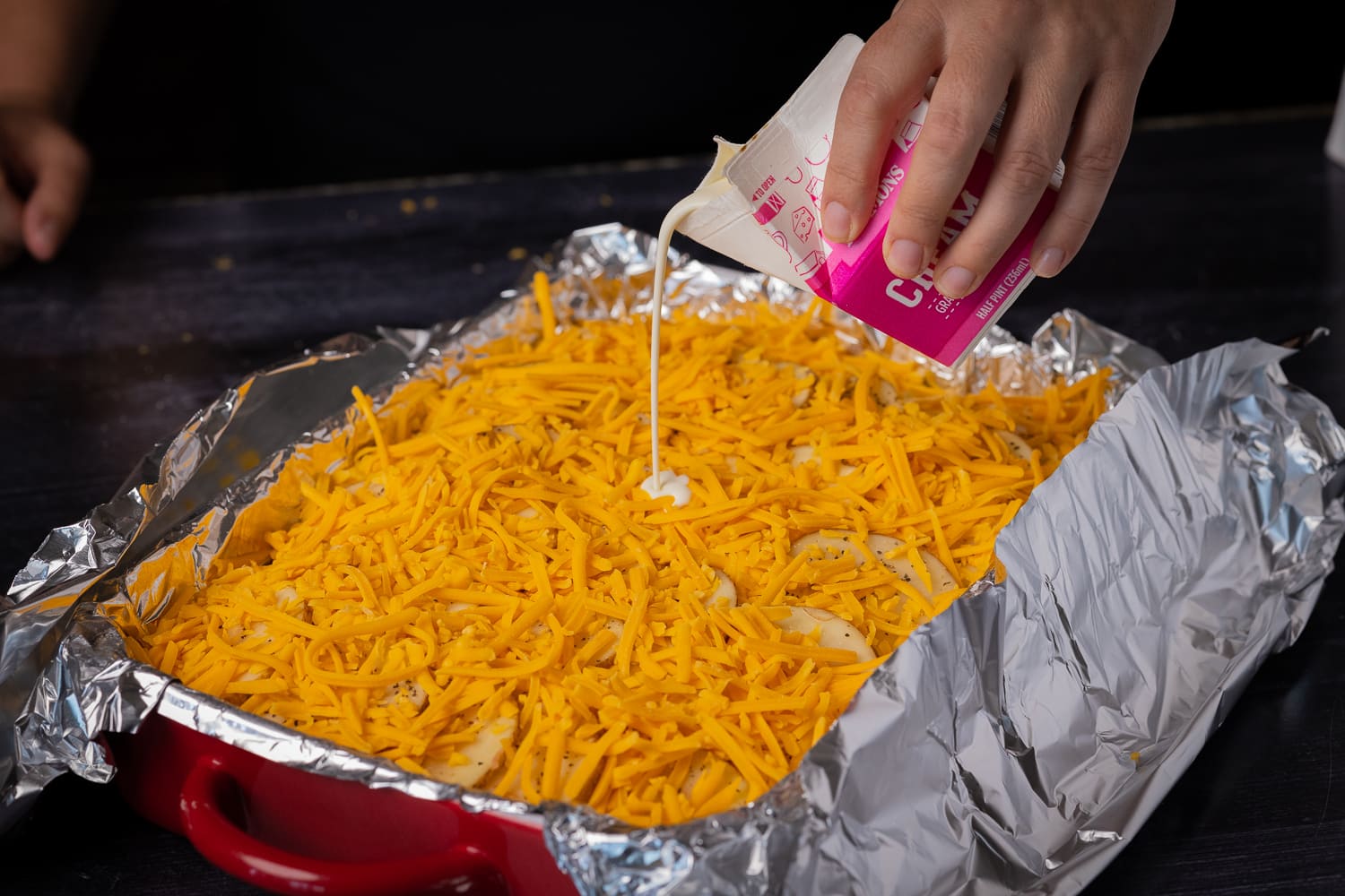 Heavy cream being poured over layers of sliced potatoes and shredded cheddar cheese.
