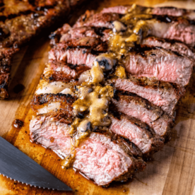 Sliced grilled rib eye steak topped with roasted garlic resting butter on a wooden cutting board.