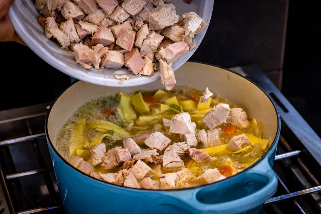 Chopped chicken being poured into a large pot of turkey noodle soup.