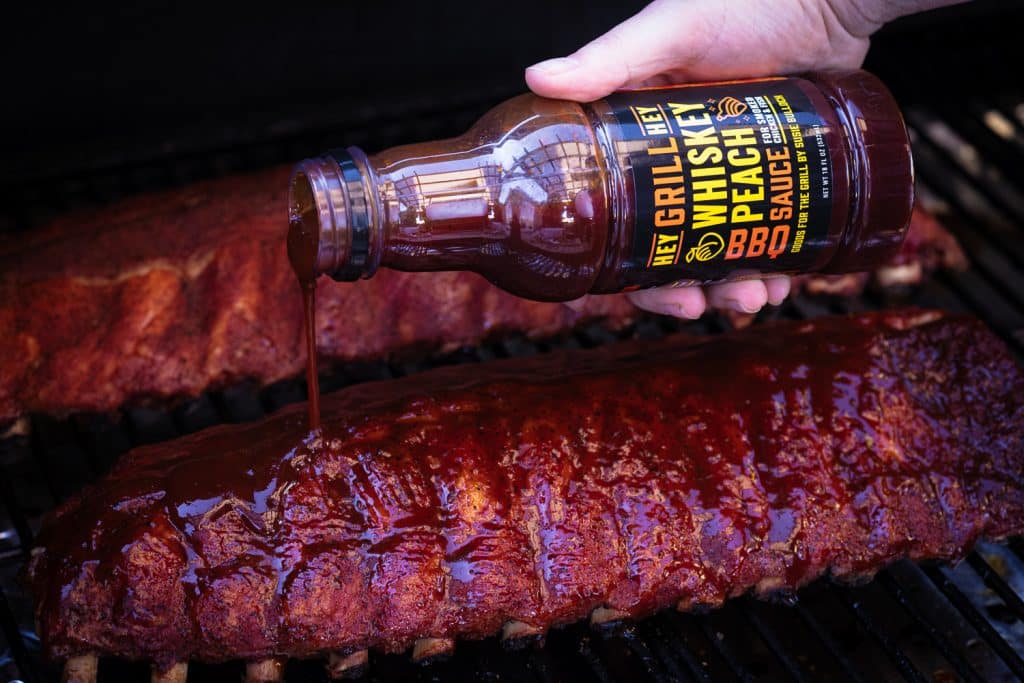 Hey Grill Hey Whiskey Peach BBQ Sauce being drizzled on a rack of baby back ribs.