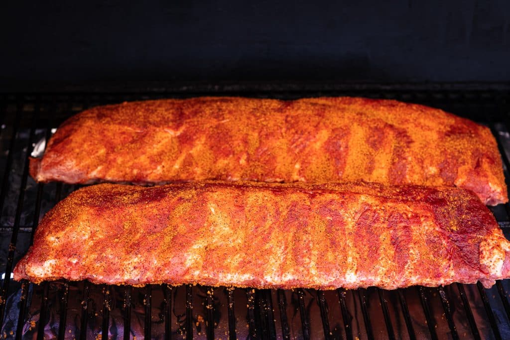 Two seasoned racks of baby back ribs on the grill grates of a smoker.