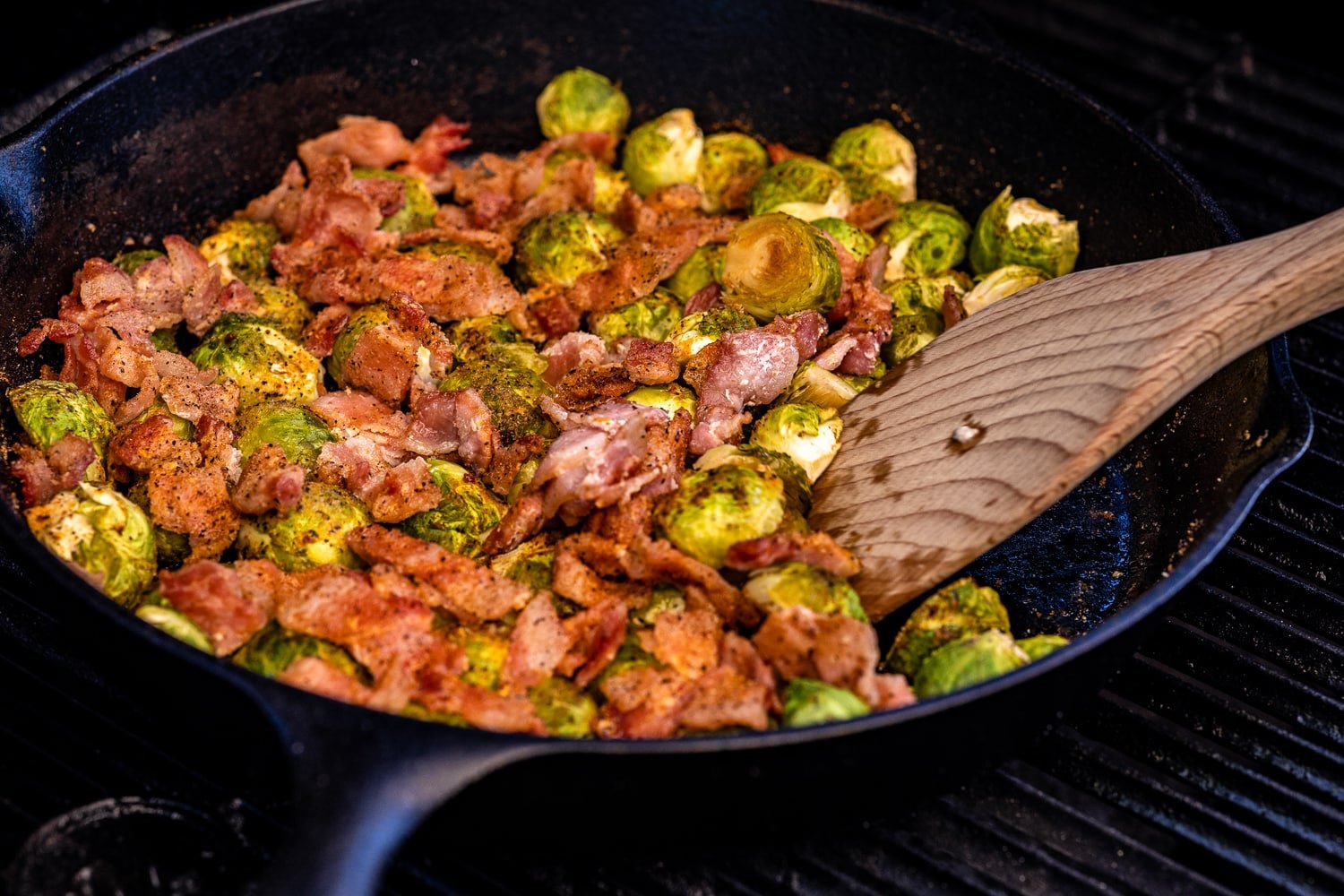 Flat wooden spoon scooping into a skillet full of seasoned bacon and Brussels sprouts.