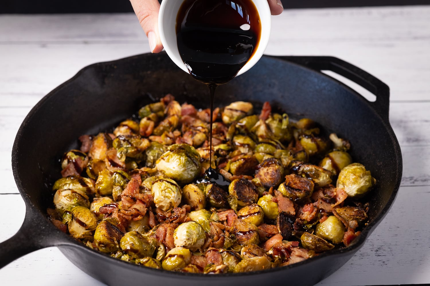 Balsamic glaze being poured into a skillet of smoked Brussels sprouts and bacon.