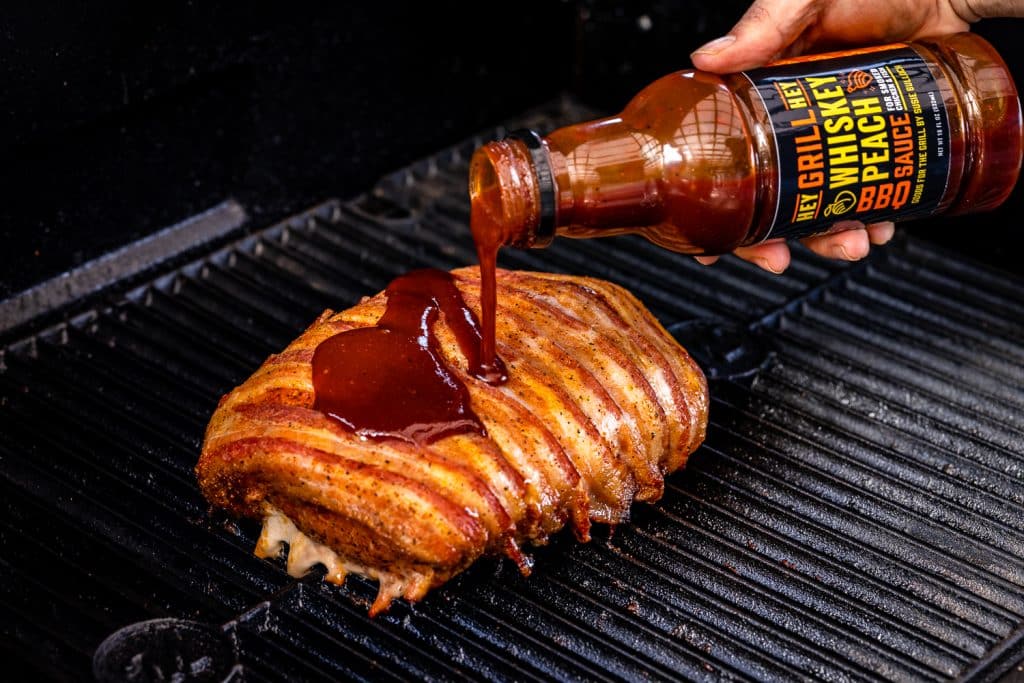 Bottle of Hey Grill Hey Whiskey Peach BBQ Sauce drizzling over a bacon wrapped pork loin on the smoker.