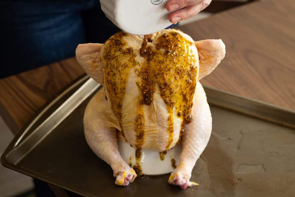 Olive oil and chicken seasoning being poured over a whole chicken on a chicken roaster.
