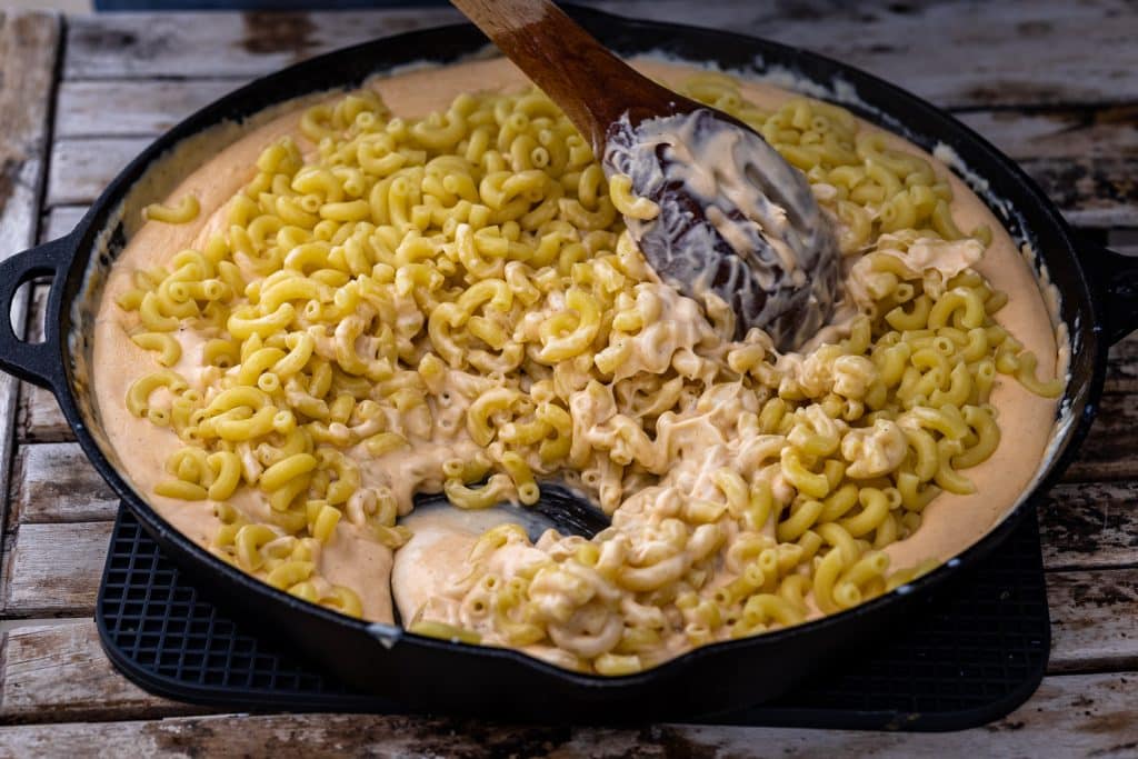 Macaroni noodles being stirred into a cast iron skillet full of cheese sauce.
