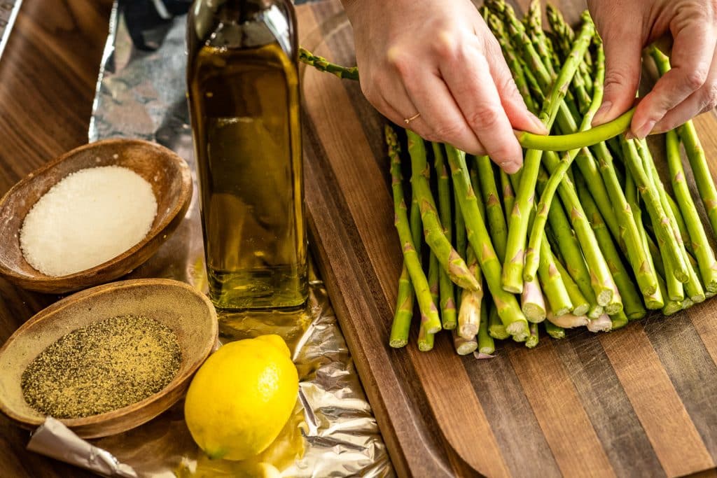 Trimmed asparagus on a wooden cutting board next to salt pepper, olive oil, and lemon.
