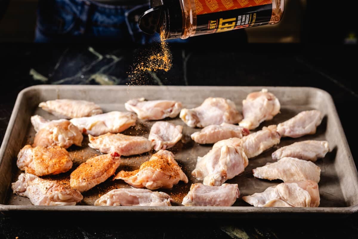 A bottle of rub being sprinkled onto raw poultry on a baking sheet below.