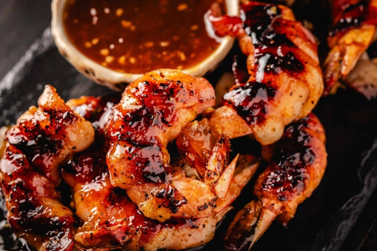 A plated of grilled shrimp wrapped in bacon next to a bowl of dipping sauce.