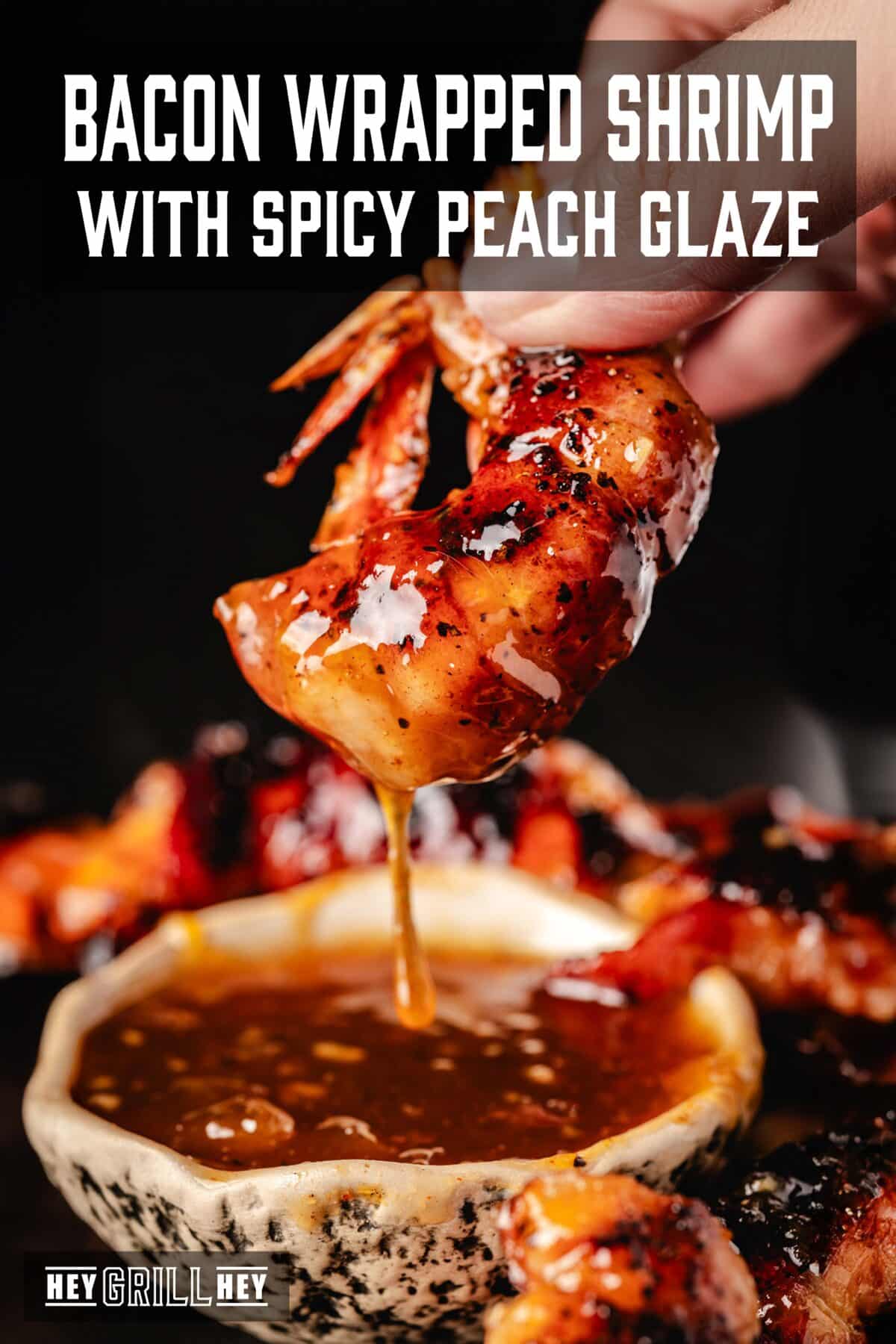 A hand holding a shrimp dripping with sauce from a bowl below. The text overlay reads "Bacon Wrapped Shrimp with Spicy Peach Glaze" at the top, and "Hey Grill Hey" at the bottom.