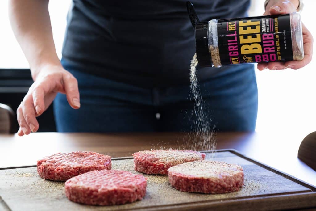 Hey Grill Hey Beef Rub being sprinkled on four hamburger patties.