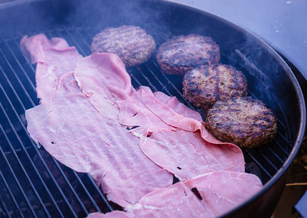Sliced corned beef and four hamburger patties on a charcoal grill.