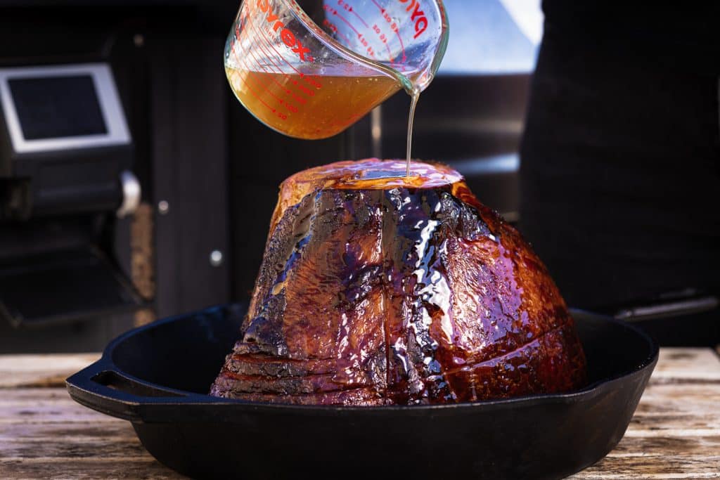Braise being poured on a spiral cut ham in a cast iron skillet.