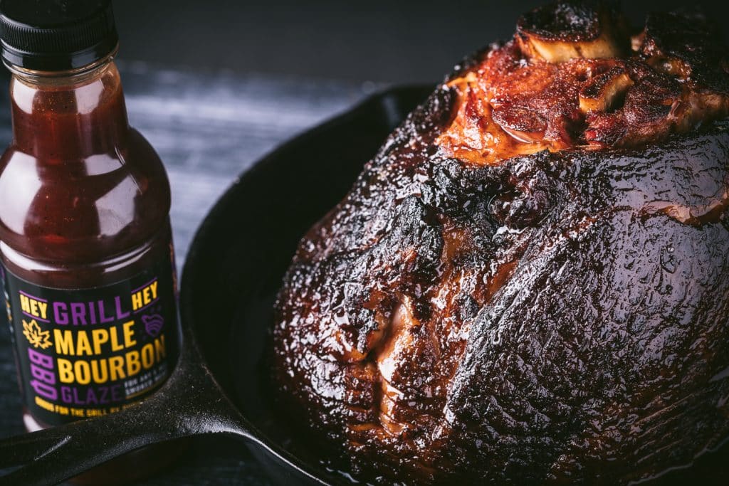 Smoked spiral ham in a cast iron skillet next to a bottle of Hey Grill Hey Maple Bourbon Grilling Glaze.