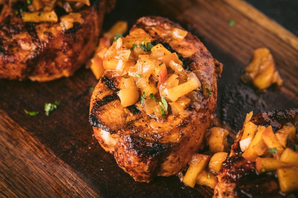 Pork loin chop topped with grilled apples and onions on a wooden cutting board.
