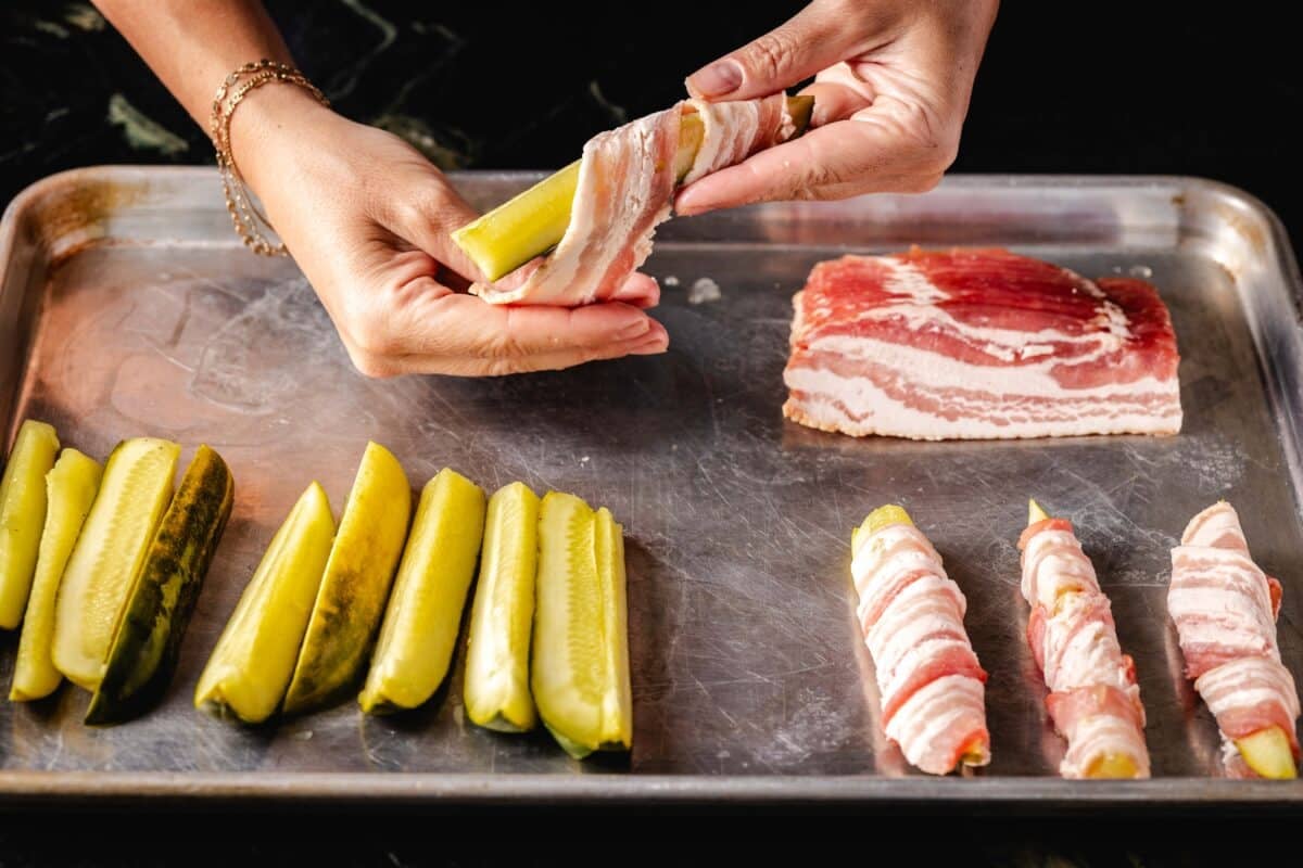 A chef's hands wrapping a dill spear with bacon above a baking sheet lined with more wrapped and unwrapped dill spears.