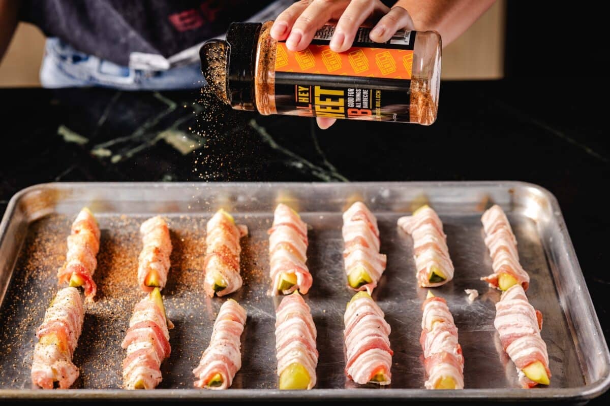 A chef's hand sprinkling a BBQ rub onto a baking sheet lined with dill spears wrapped in bacon.