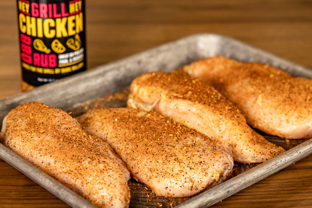 Four chicken breasts seasoned with Hey Grill Hey Chicken Rub on a metal baking dish.