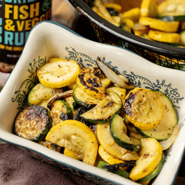 Grilled zucchini and squash in a serving dish next to a bottle of Hey Grill Hey Fish & Veggie Rub.