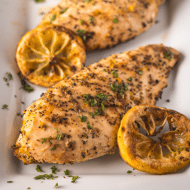 Two grilled lemon pepper chicken breasts on a platter next to grilled lemon slices.