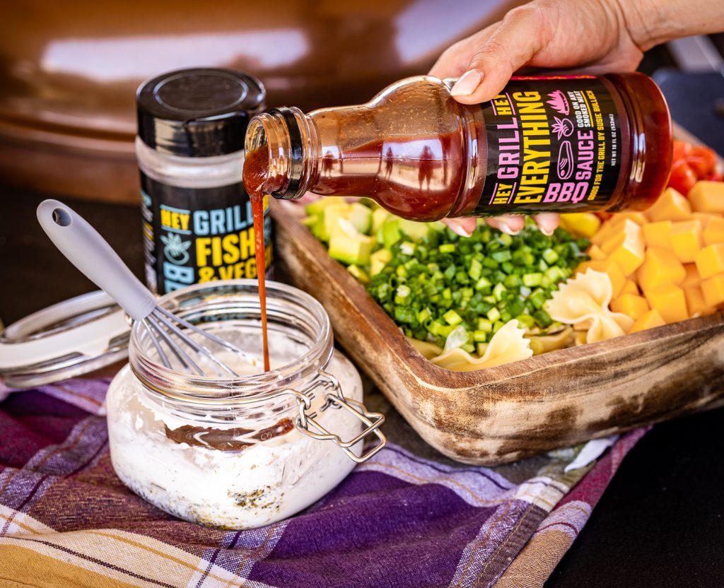 Hey Grill Hey Everything BBQ sauce being added to BBQ ranch dressing next to a bowl of pasta salad.
