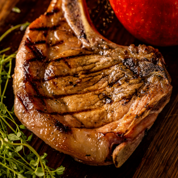Brined and grilled pork chop on a cutting board next to herbs and apples.