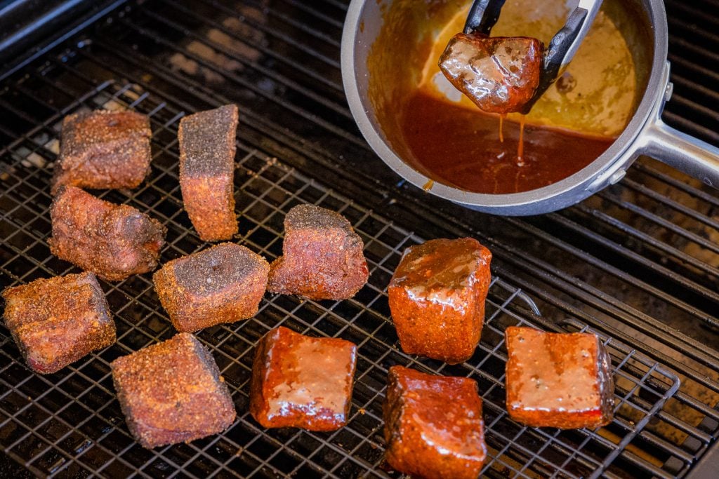 Steak bites being dipped into BBQ sauce and placed back on the grill grates of the smoker.