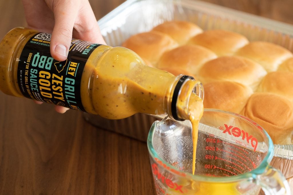 Zesty Gold BBQ Sauce being poured into a glass measuring cup next to a baking pan of Hawaiian rolls.