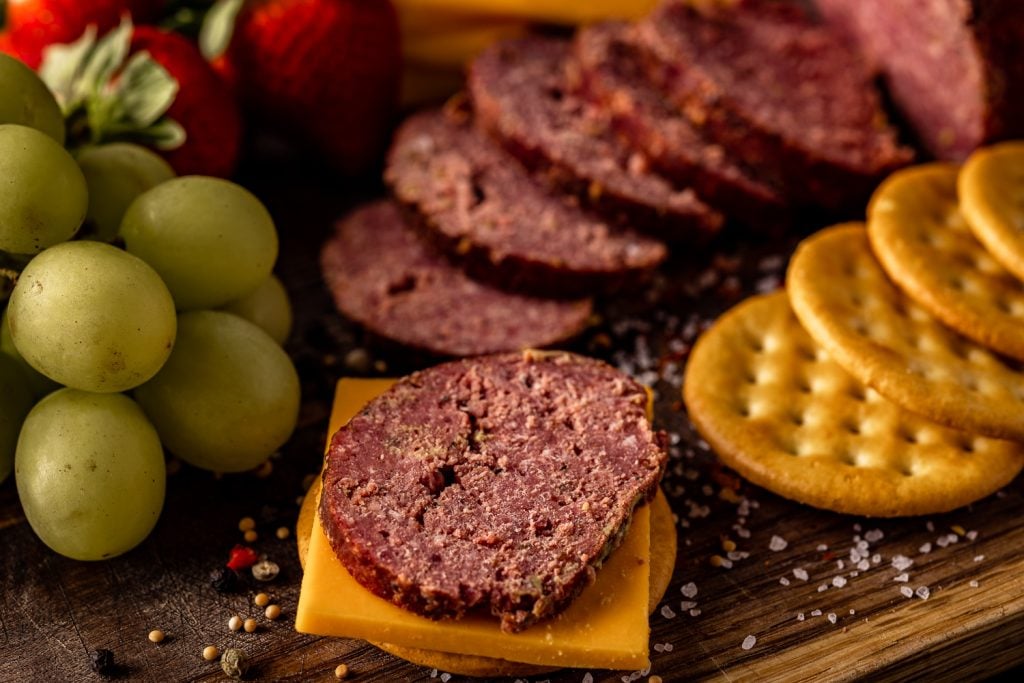 Sliced smoked summer sausage on a cutting board with crackers, grapes, and fruit.