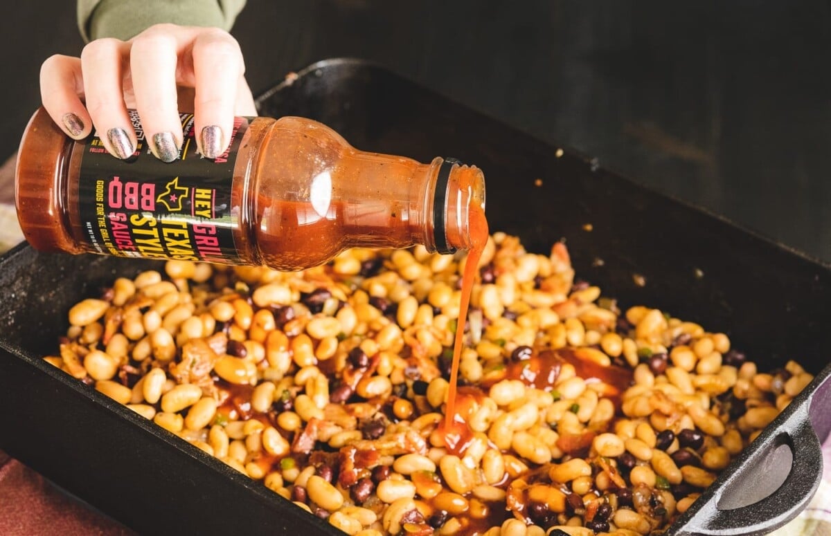 Texas BBQ sauce being added to a pan of BBQ baked beans on the grill.