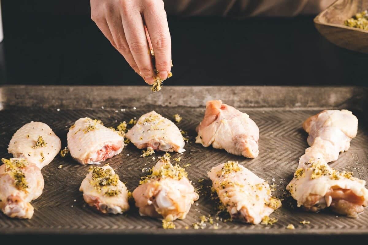 Chicken wings on baking sheet being sprinkled with lemon zest and salt and pepper seasoning.