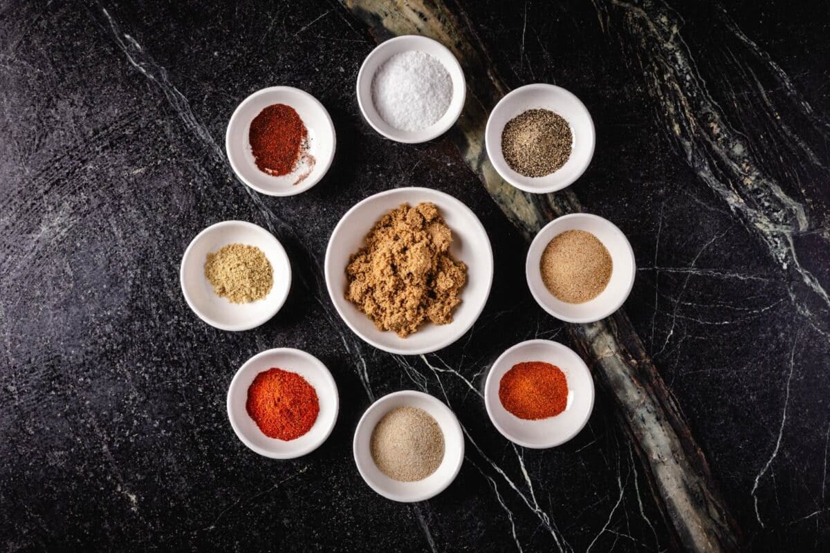 A large glass bowl with brown sugar surrounded by 8 smaller bowls of seasonings and spices.