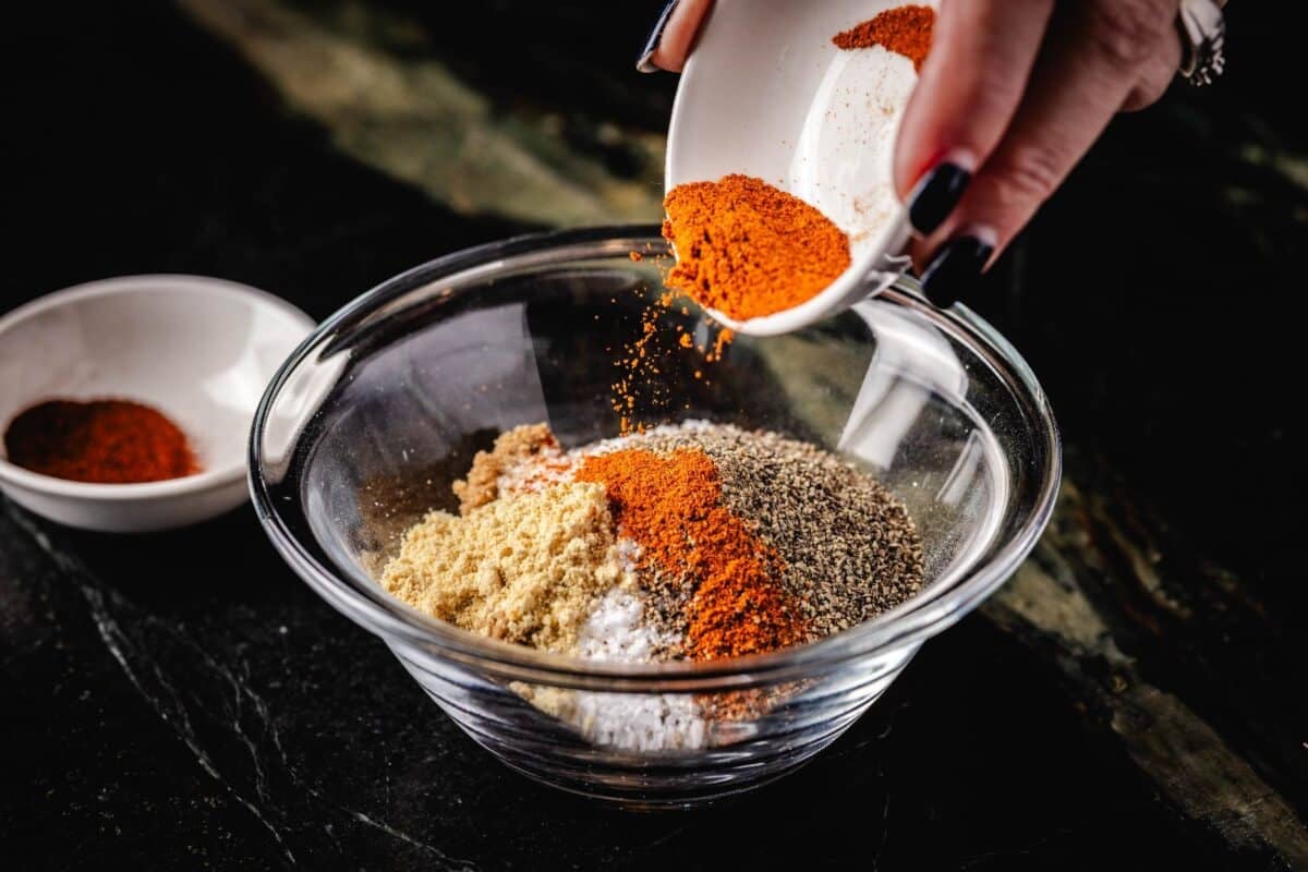 Cayenne in a white bowl being poured into a larger bowl full of various spices.