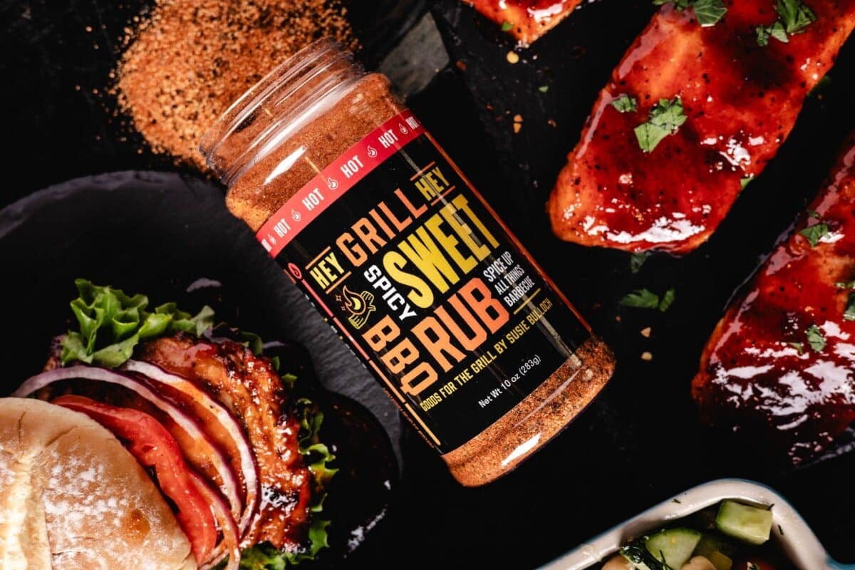 A jar of Spicy Sweet Rub lying on a surface spilling out next to grilled veggies.