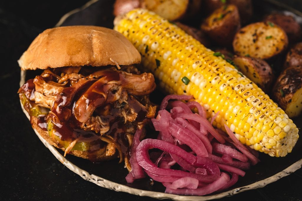 BBQ pulled chicken sandwich next to corn on the cob.
