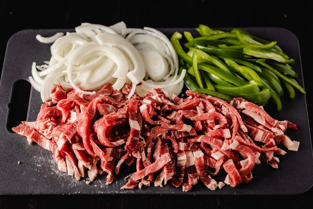 Sliced ribeye steak, onions, and green peppers on a cutting board.