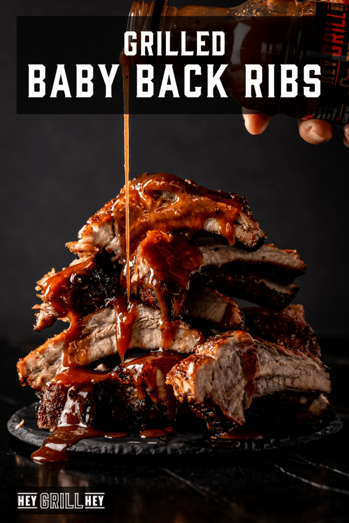 Pile of grilled baby back ribs being drizzled with BBQ sauce with text overlay - Grilled Baby Back Ribs.