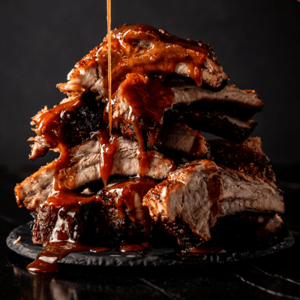 Pile of grilled baby back ribs being drizzled with BBQ sauce.