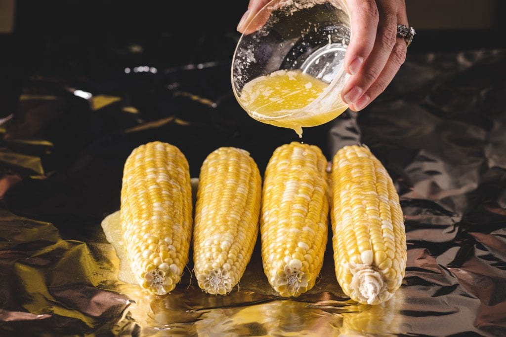 Four cobs of corn being seasoned with melted butter.