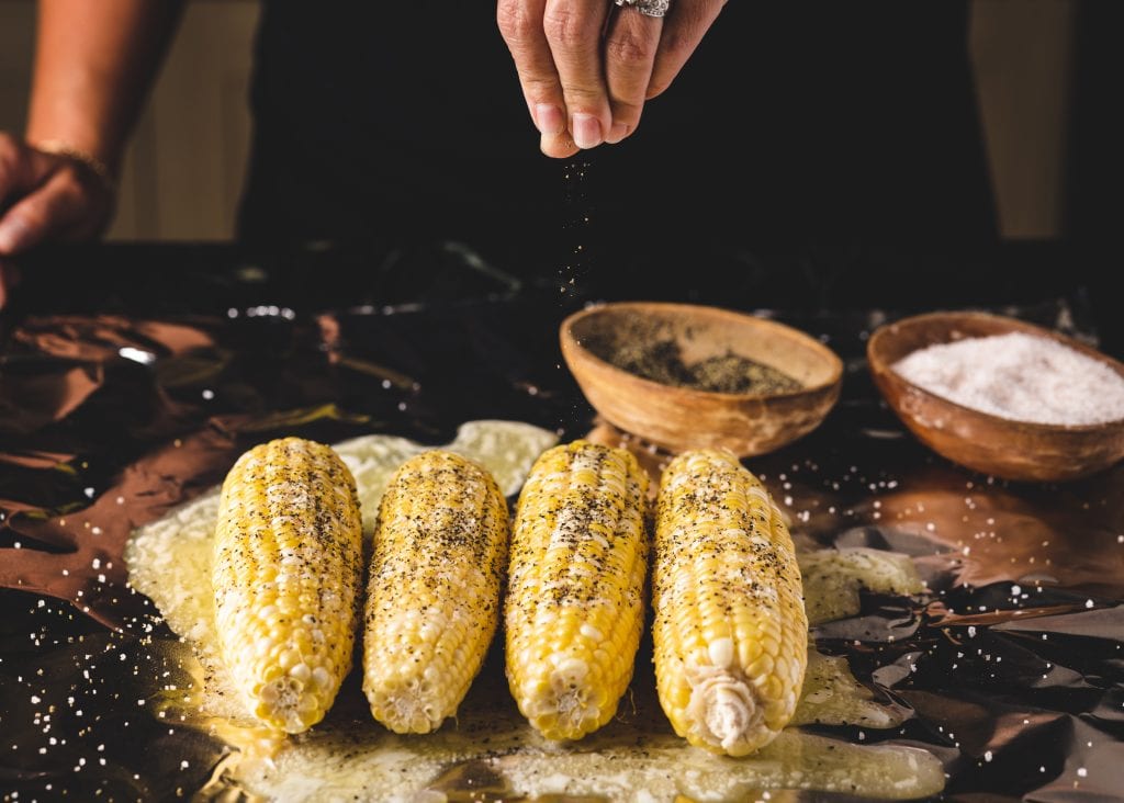 Four cobs of corn being seasoned with black pepper.