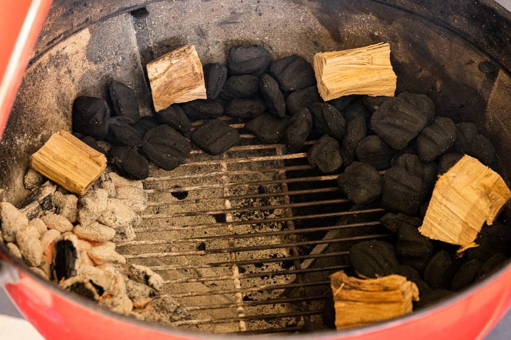 Charcoal is arranged around the charcoal chamber of a kettle grill. Unlit briquettes are topped with wood chunks.