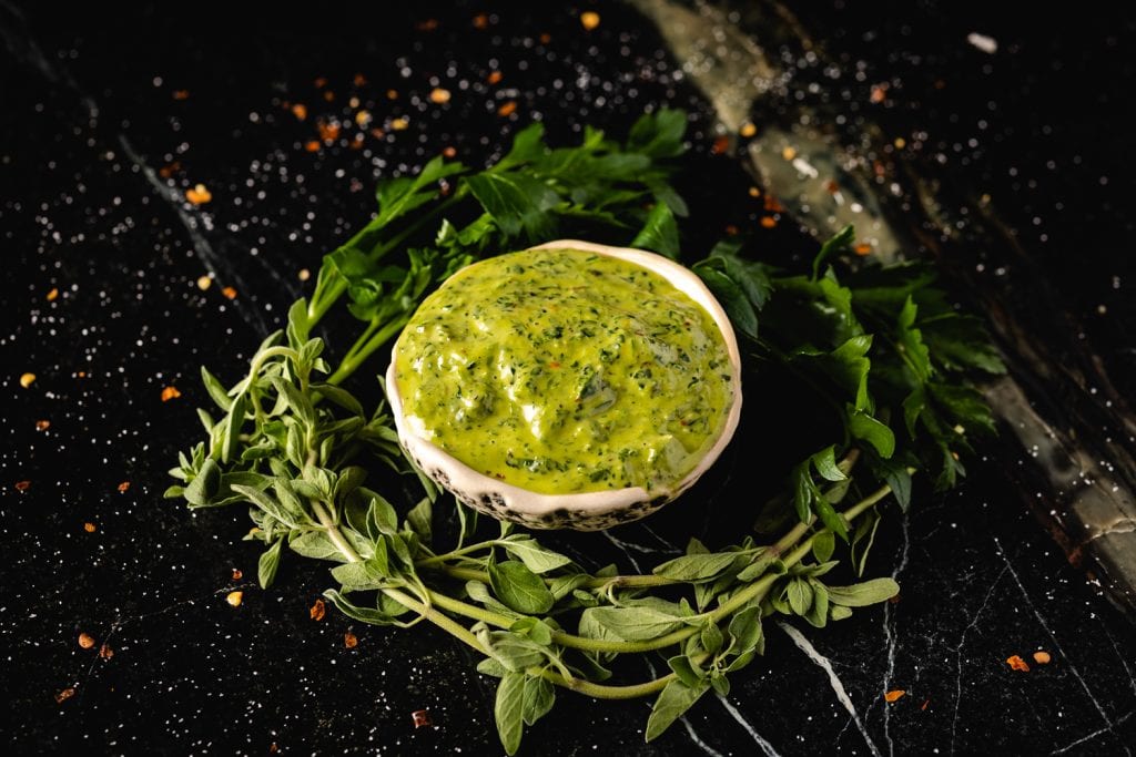 Bowl of chimichurri surrounded by herbs with text overlay - The Best Chimichurri.